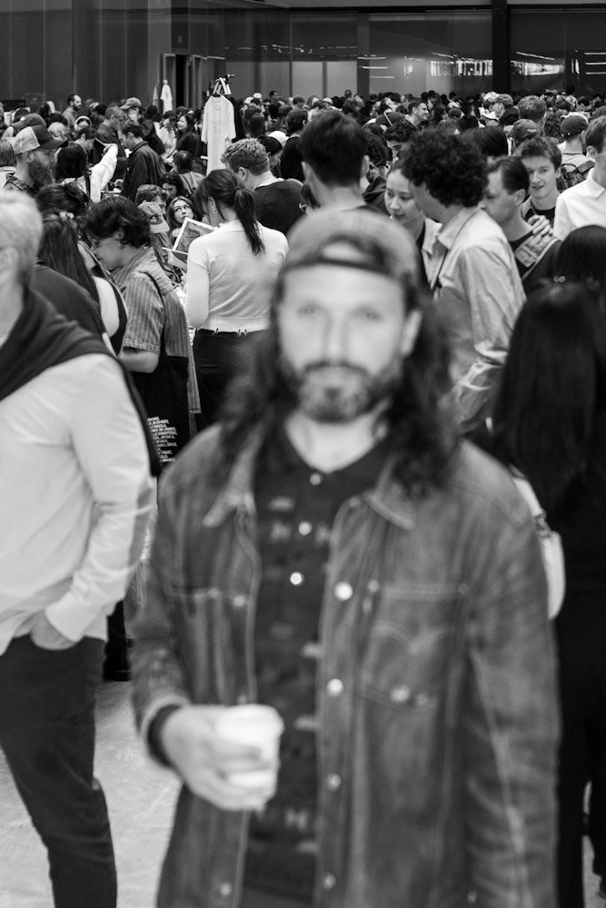 A crowded event featuring a diverse group of people, with a focus on a man in the foreground holding a coffee cup, dressed in a denim jacket.