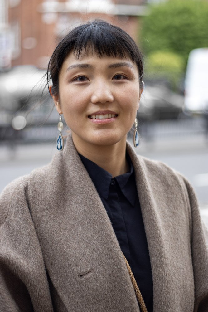Portrait of a smiling woman with dark hair wearing earrings and a brown coat, standing outdoors in an urban setting.